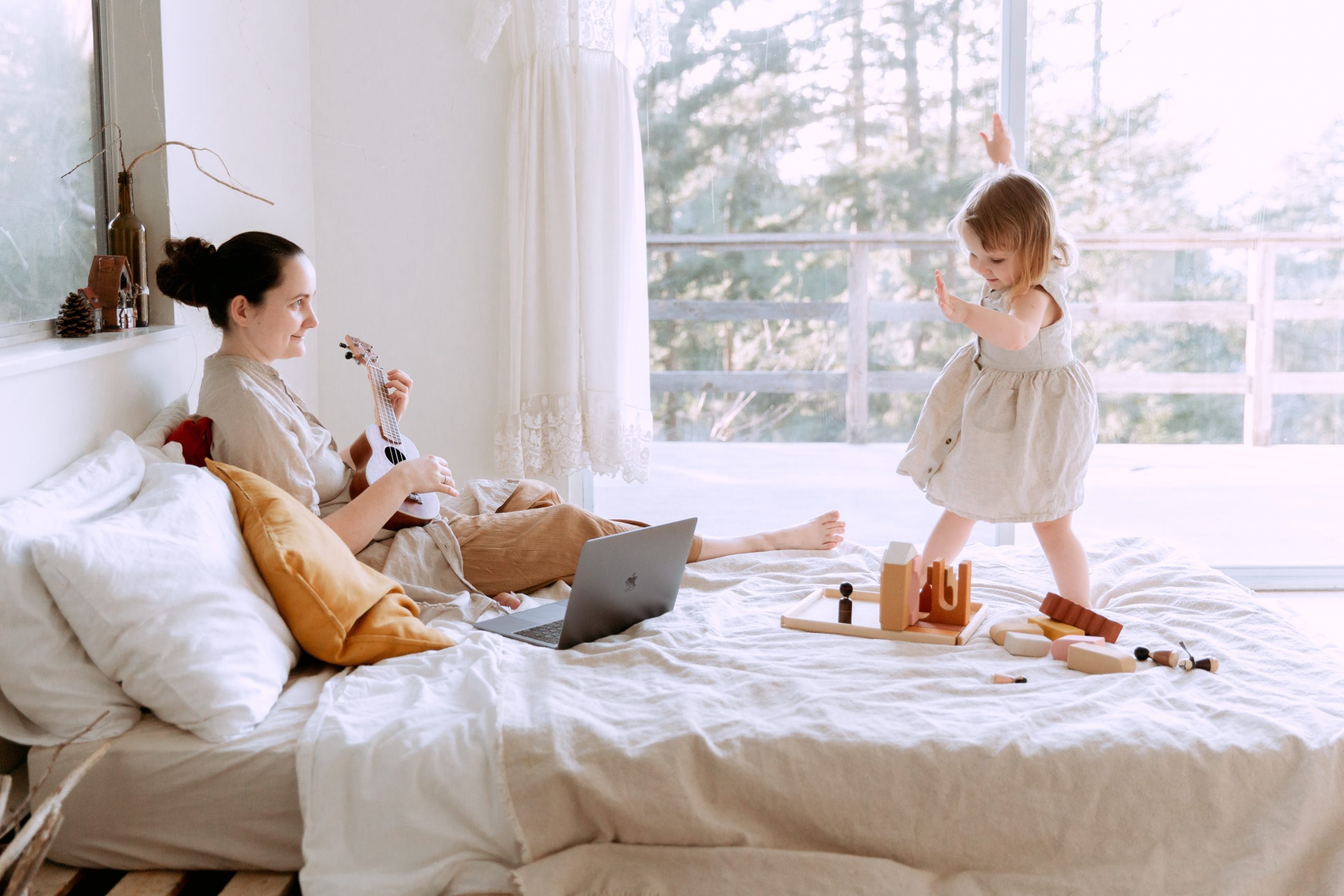 Petite fille qui joue sur le lit avec sa maman qui joue d'un instrument.